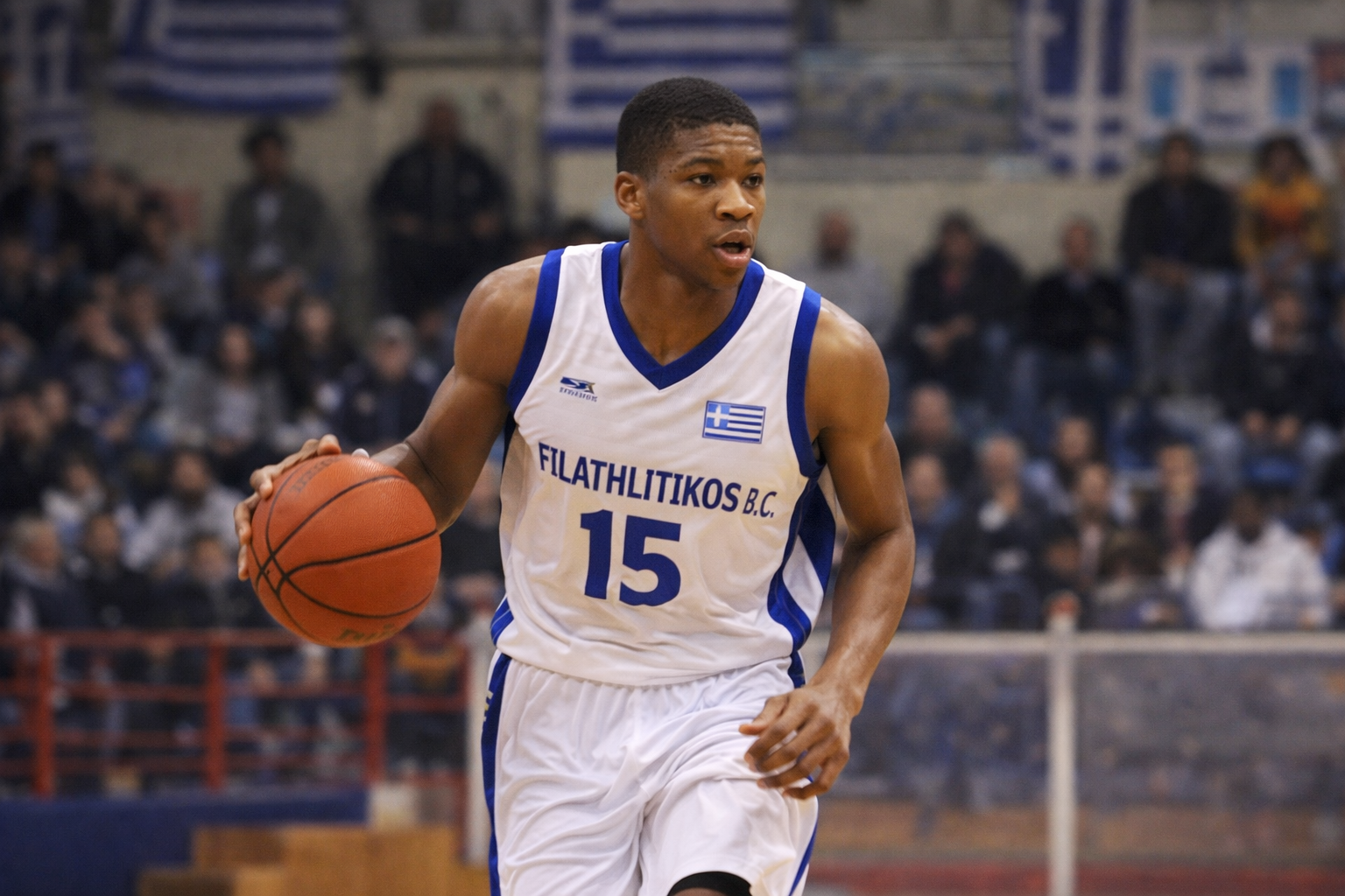 Foto Giannis Antetokounmpo muda tahun 2013 saat bermain untuk Filathlitikos B.C. di Yunani, mengenakan jersey putih dan biru nomor 15, dribel bola basket dengan fokus tinggi di lapangan gymnasium yang dipenuhi penonton dan bendera Yunani di latar belakang.