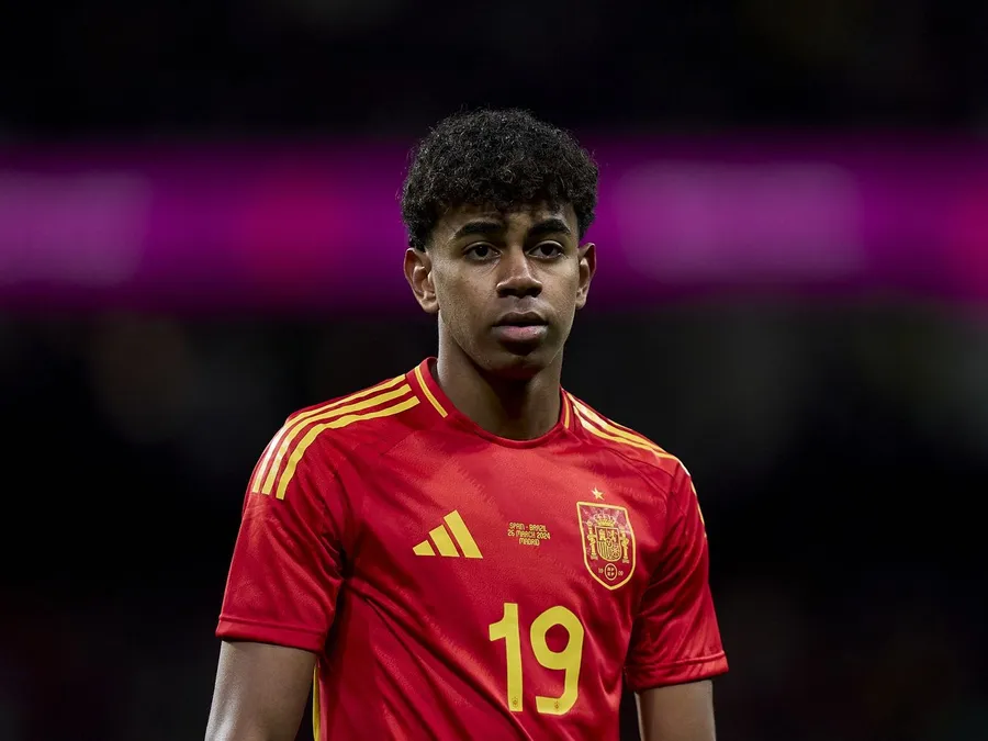 Seorang pemain sepak bola muda mengenakan jersey tim nasional Spanyol berwarna merah dengan nomor punggung 19, berdiri di lapangan dengan ekspresi fokus di bawah sorotan lampu stadion. Lamine Yamal