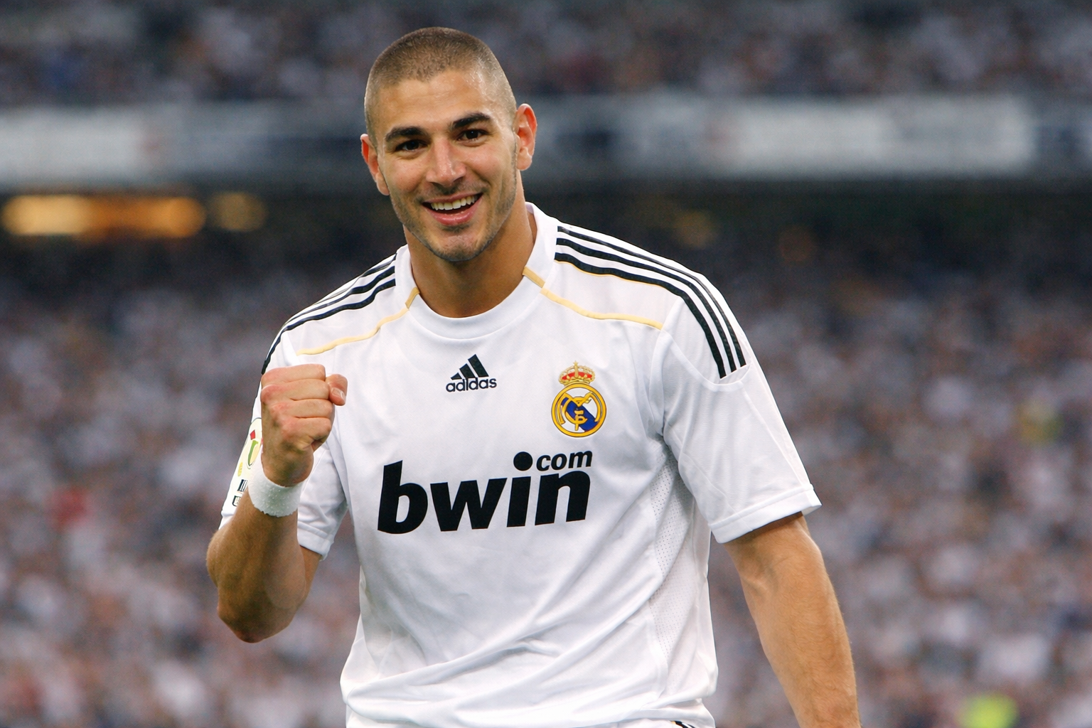 Karim Benzema mengenakan jersey putih Real Madrid musim 2009, tersenyum sambil mengepalkan tangan di lapangan stadion dengan latar belakang penonton yang buram
