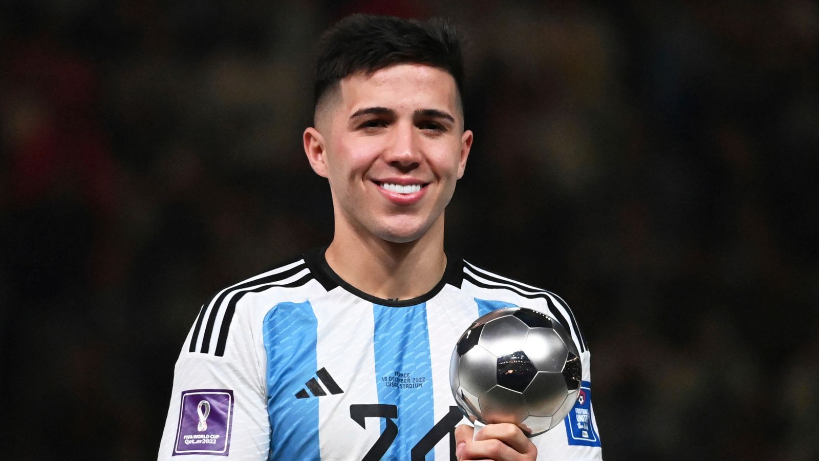 Seorang pemain sepak bola Enzo Fernandez tim nasional Argentina mengenakan jersey biru-putih sambil memegang trofi individu di stadion dengan latar belakang gelap.